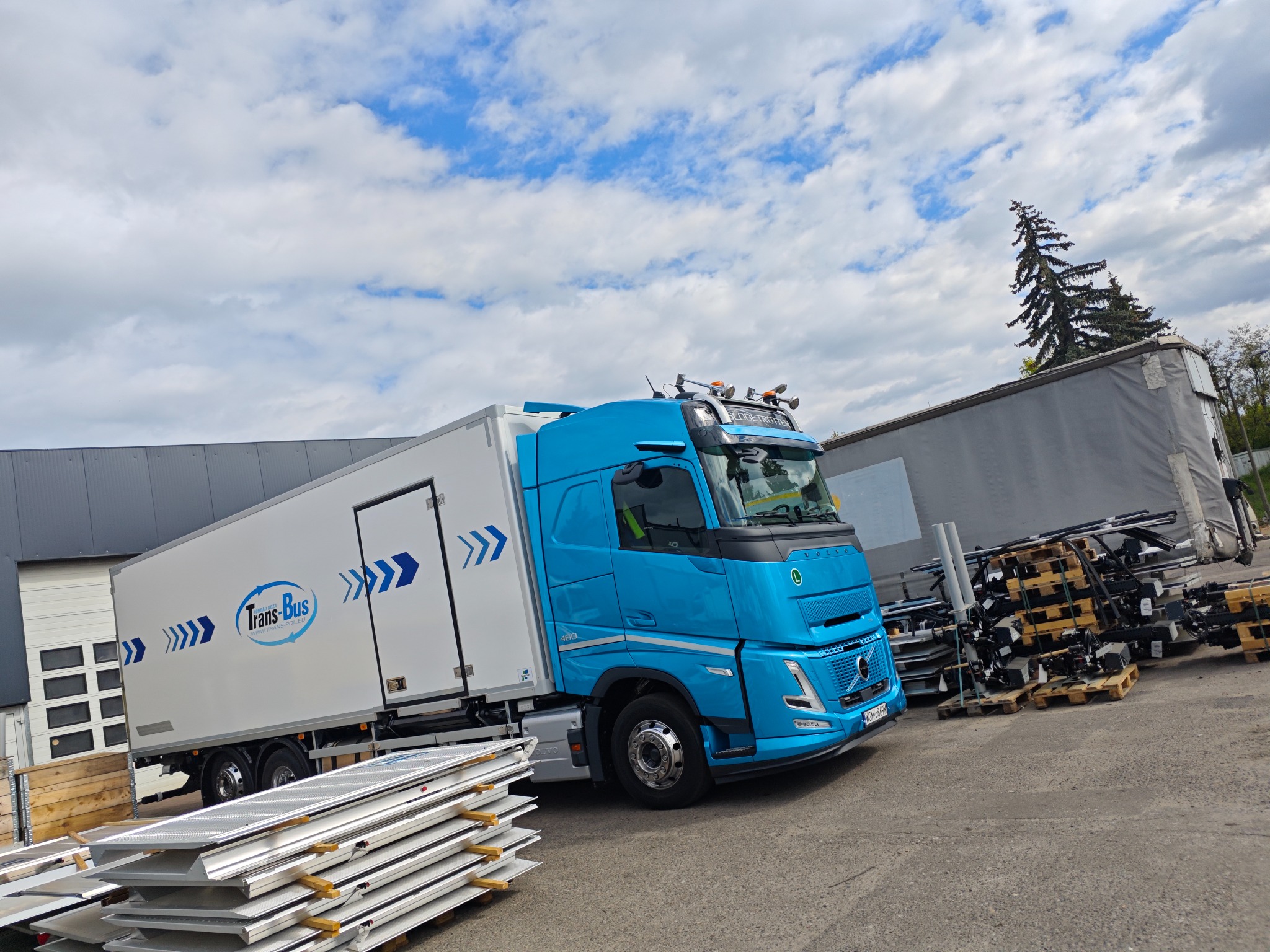 Błękitny bus z naczepą Trans-Bus na placu, obok składowane panele i palety. Volvo 460 z charakterystycznym logo firmy. Błękitne niebo z chmurami w tle.