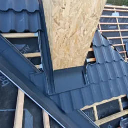 Detal montażu czarnej blachodachówki wokół komina z desek. Widoczne łaty, membrana dachowa i obróbki blacharskie. Kątowe ujęcie z góry, w świetle dziennym.