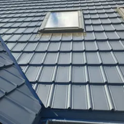 Szary dach z dachówki z oknem dachowym, widok z dołu. Elementy blacharskie i rynny w kolorze dachu. Fragment ściany budynku w tle. Dachówka karpiówka.