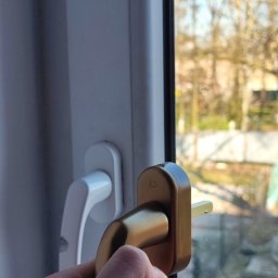 SerwisTIM - Dłoń przytrzymuje złotą klamkę okna PCV na tle szyby i białej ramy, z widokiem na zewnątrz. Ostrość na detale klamki i mechanizm zamka.