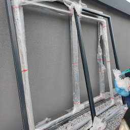 SerwisTIM - Nowe ramy okienne PCV w folii ochronnej, przygotowane do montażu w Zabrzu. Szare ramy opierają się o ścianę, widoczne elementy montażowe.