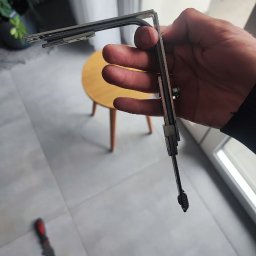 SerwisTIM - Ręka trzyma uszczelkę okienną z profilem metalowym na tle jasnej podłogi i stołka. Widoczne narzędzia sugerują naprawę lub wymianę uszczelki.