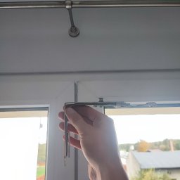SerwisTIM - Ręka trzyma metalowy element mechanizmu rolety okiennej na tle okna PCV, widoczny fragment karnisza. Drobiazgowa naprawa z dbałością o detale.