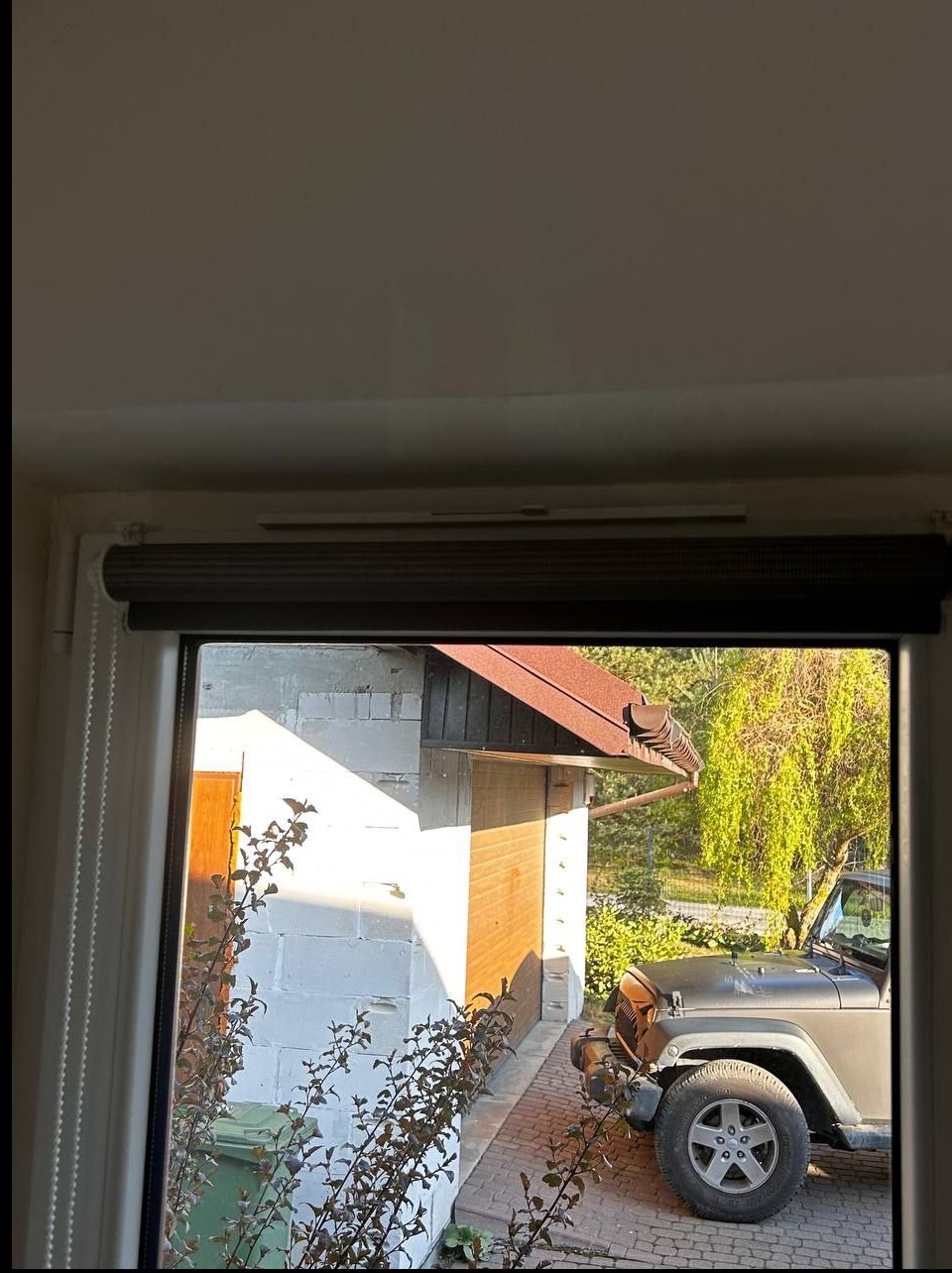 Zamknięta, szara roleta okienna z widokiem na budynek gospodarczy z brązowym dachem i szarym samochodem na podjeździe w słoneczny dzień.