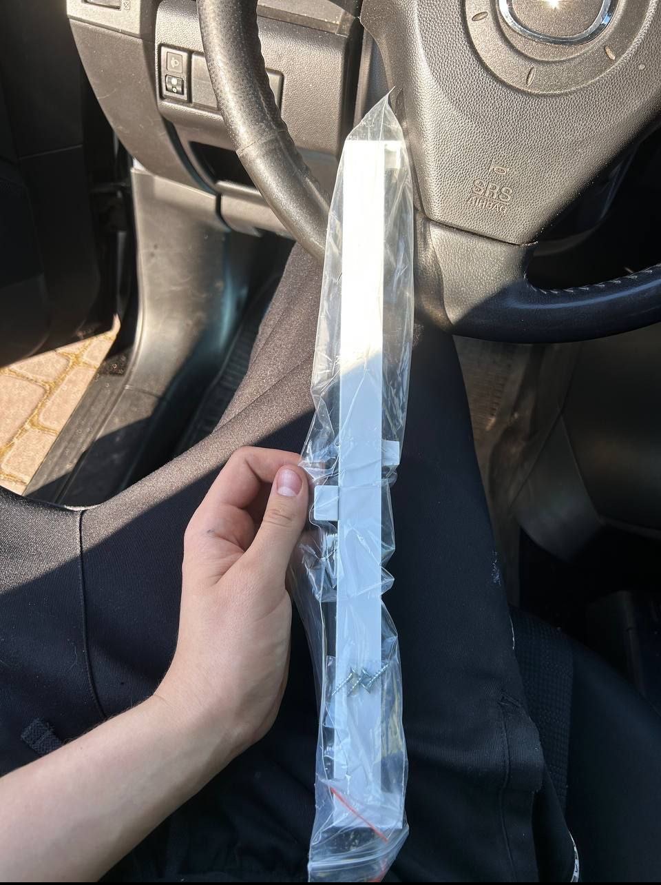 Biała listwa okienna PCV w folii, trzymana w dłoni na tle deski rozdzielczej samochodu. Widoczne śruby montażowe w opakowaniu.
