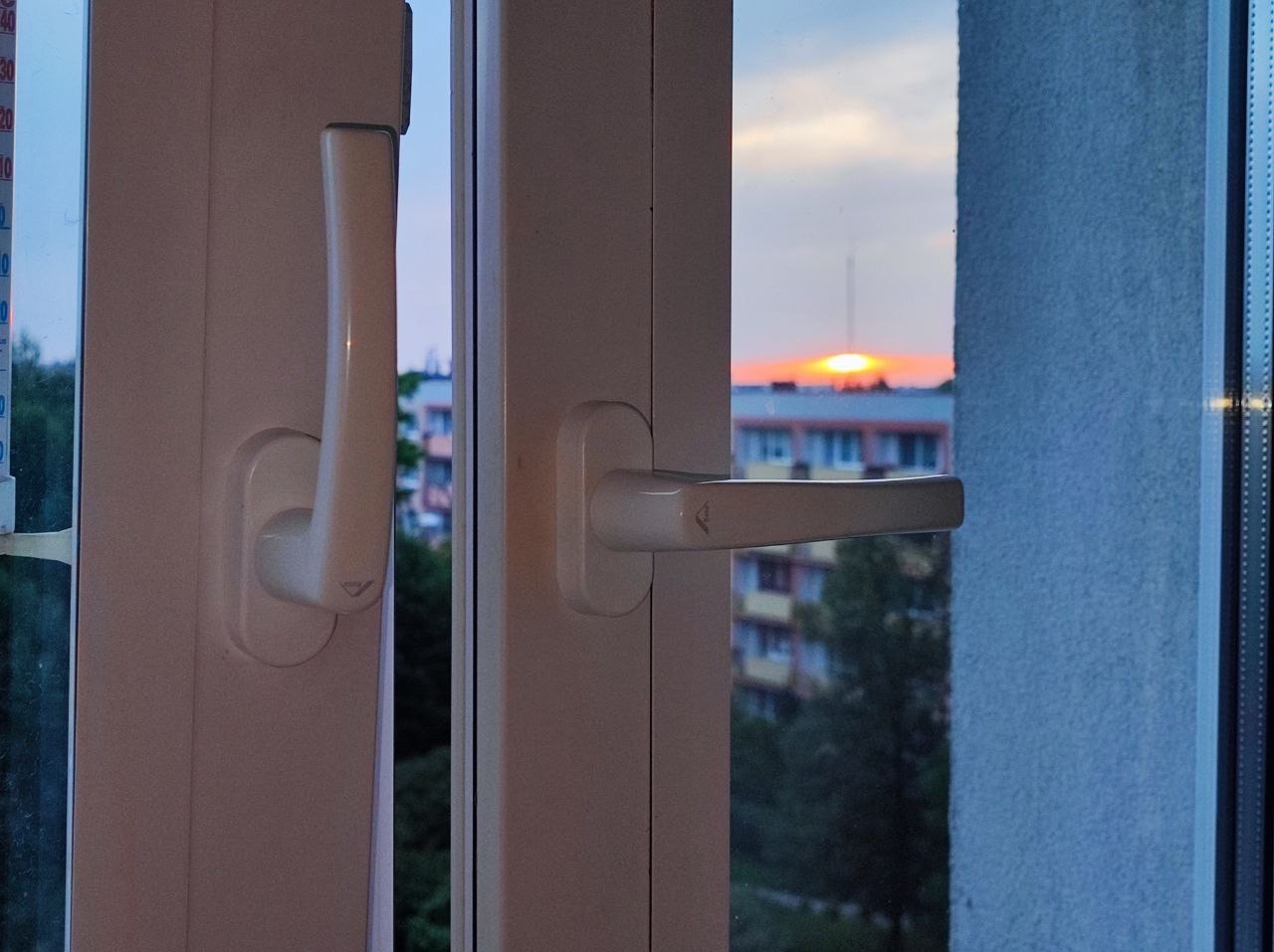 Białe okno PCV z widokiem na zachodzące słońce i blok mieszkalny. Termometr przymocowany do ramy okiennej. Widoczne dwie klamki okienne.