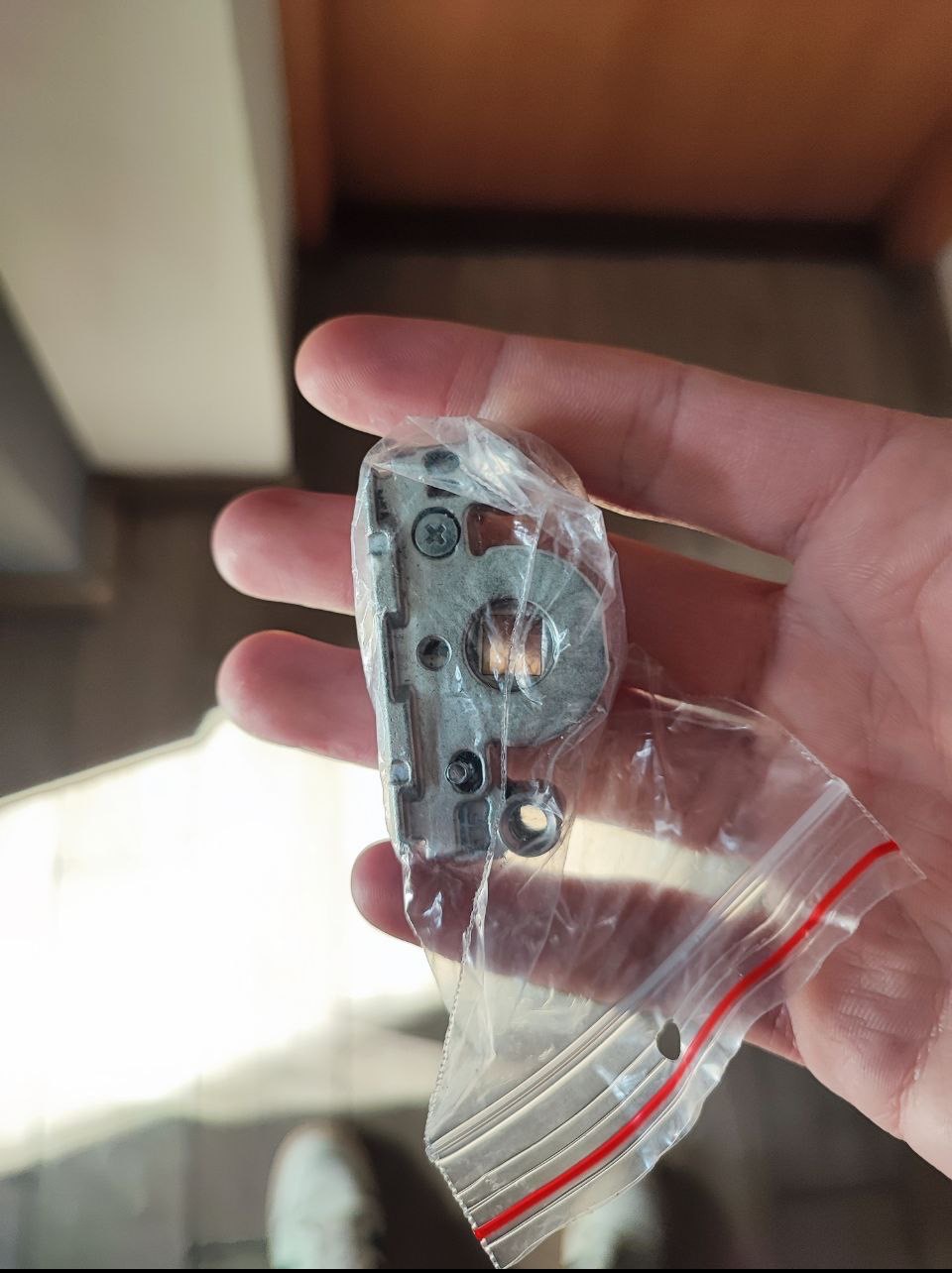 Dłoń trzyma w woreczku zasuwnicę okienną z widocznymi śrubami, element okucia okiennego do naprawy, na tle jasnego wnętrza.