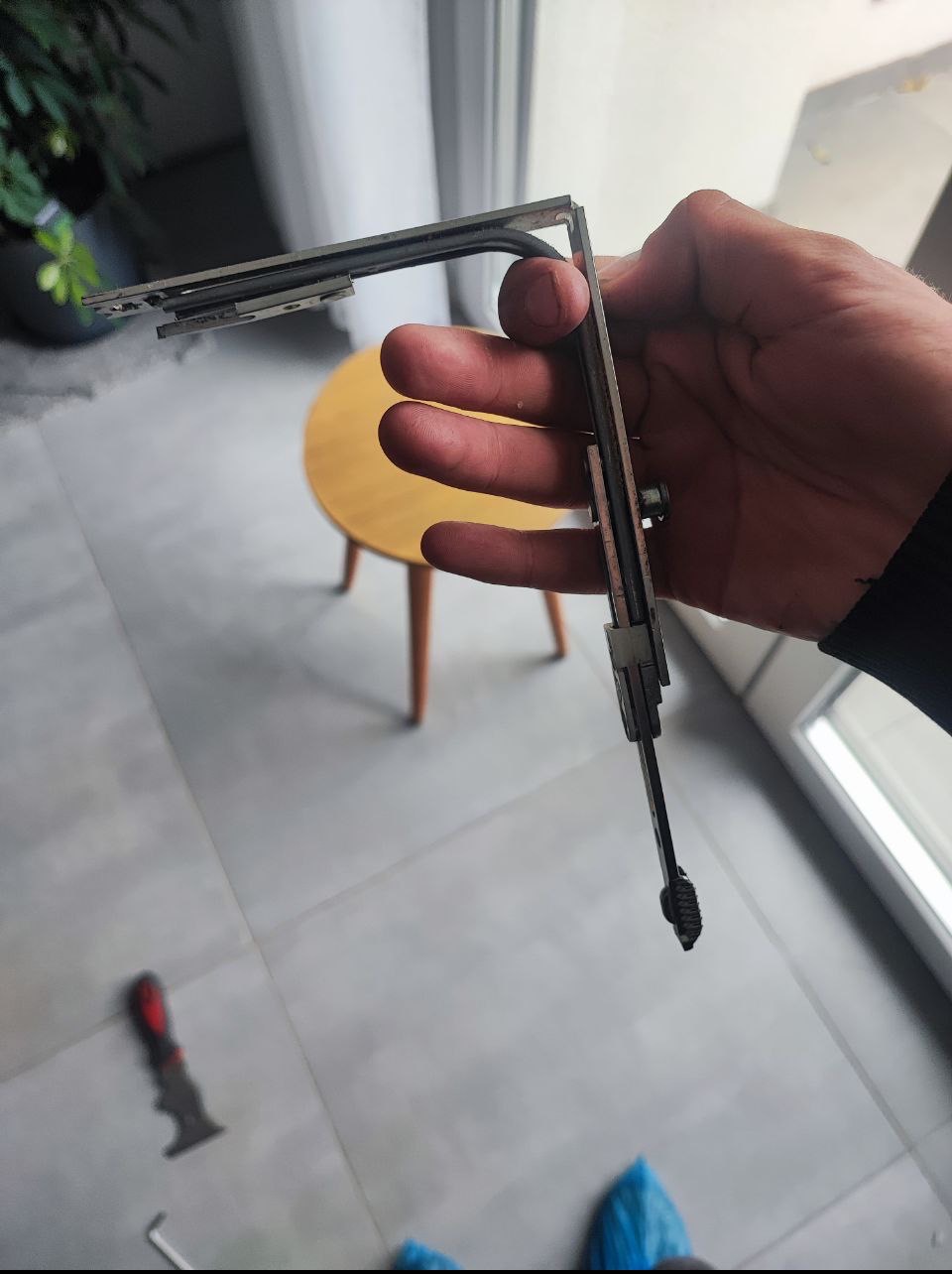 Ręka trzyma uszczelkę okienną z profilem metalowym na tle jasnej podłogi i stołka. Widoczne narzędzia sugerują naprawę lub wymianę uszczelki.