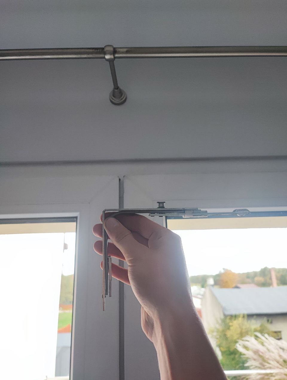 Ręka trzyma metalowy element mechanizmu rolety okiennej na tle okna PCV, widoczny fragment karnisza. Drobiazgowa naprawa z dbałością o detale.