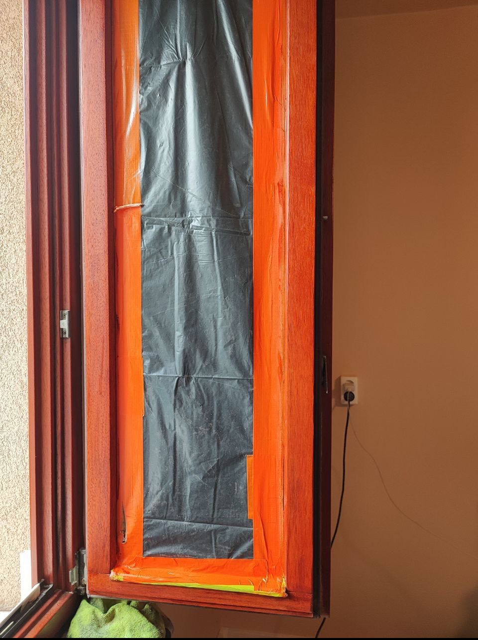 Otwarta drewniana rama okienna zabezpieczona folią i taśmą malarską przed malowaniem, widoczny fragment ściany z gniazdkiem elektrycznym.