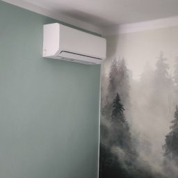 CELTERKLIM - Biały klimatyzator na ścianie w pokoju z tapetą w leśny motyw. Widok z dołu, urządzenie zamontowane blisko sufitu. Ściana pomalowana na zielono.