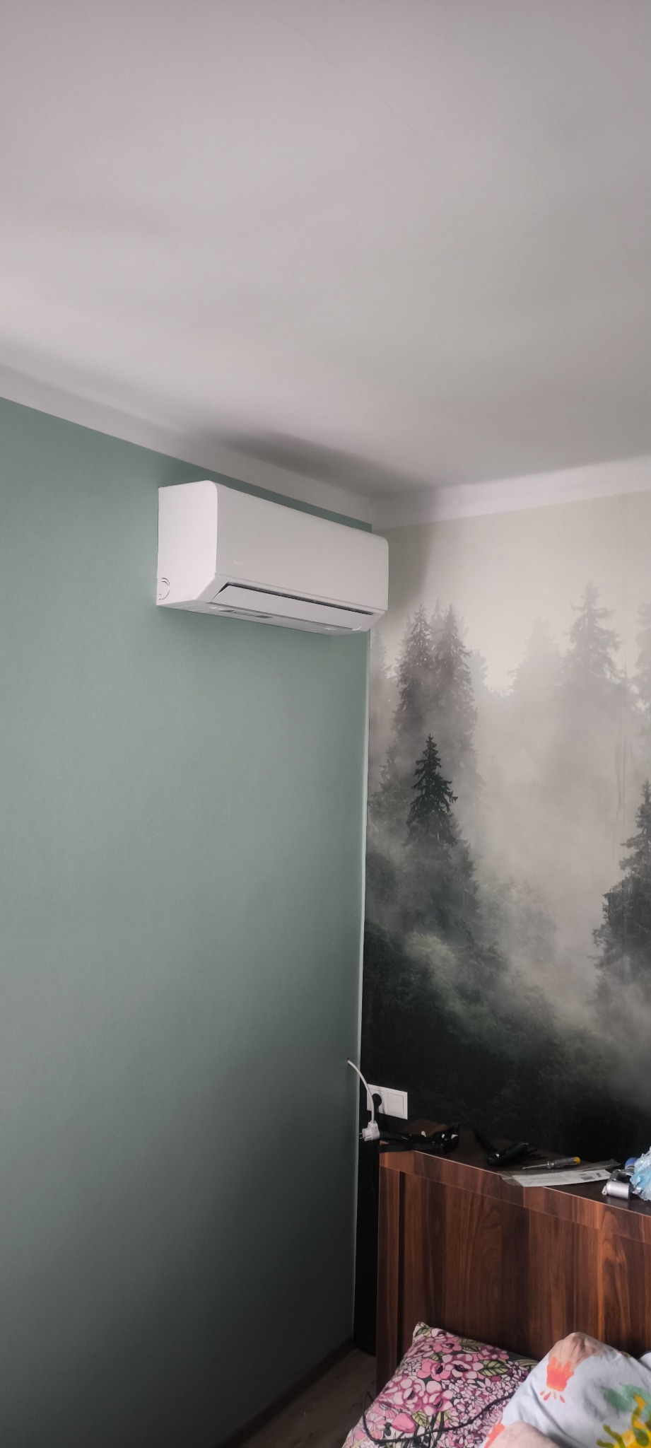 Biały klimatyzator na ścianie w pokoju z tapetą w leśny motyw. Widok z dołu, urządzenie zamontowane blisko sufitu. Ściana pomalowana na zielono.