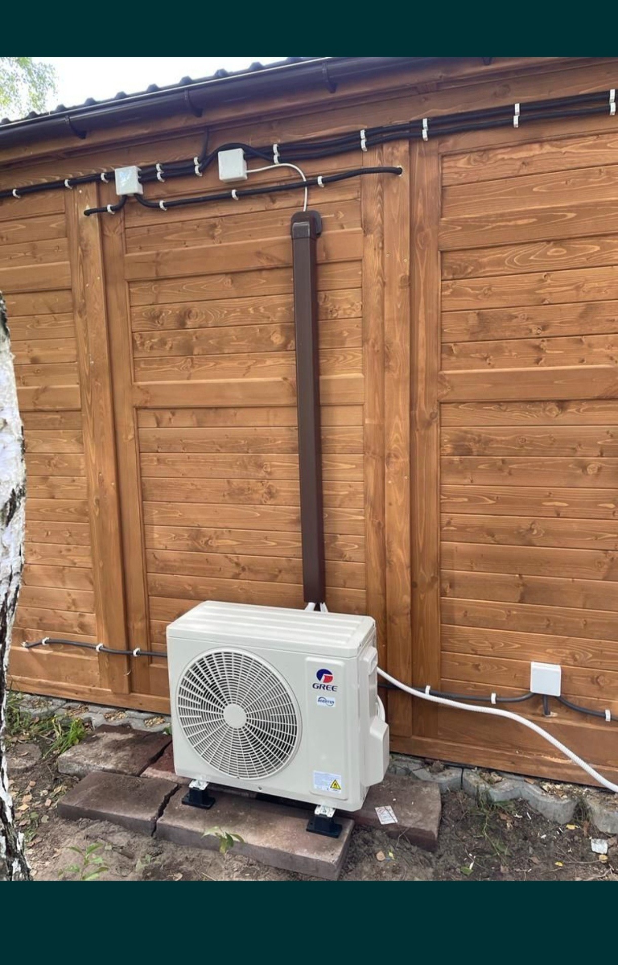Zewnętrzna jednostka klimatyzacji Gree zamontowana przy drewnianej ścianie budynku z widocznymi przewodami i puszkami instalacyjnymi.