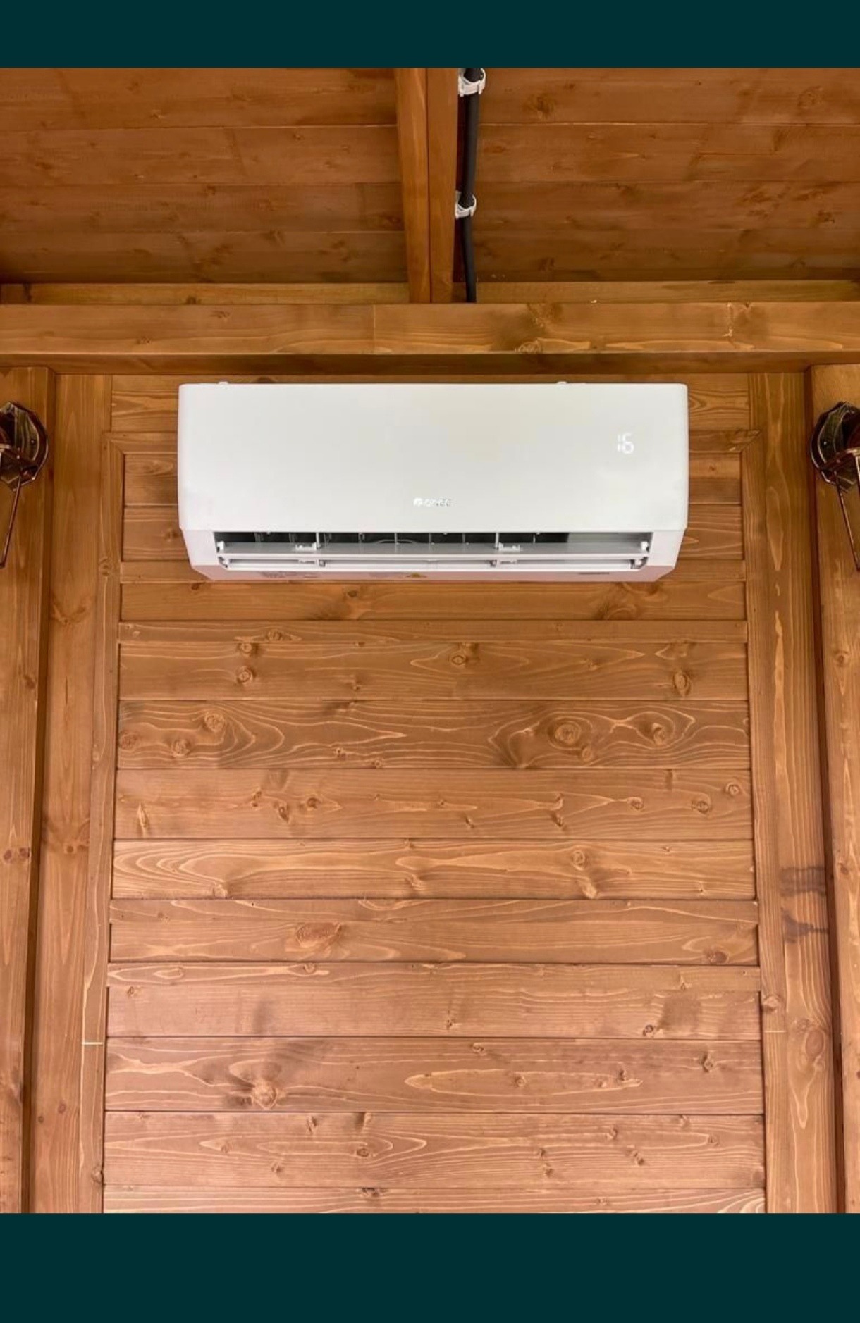 Biały klimatyzator ścienny marki Gree zamontowany na drewnianej ścianie wewnątrz pomieszczenia z drewnianym sufitem i ozdobnymi lampami po bokach.