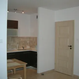 Wnętrze apartamentu: widok na aneks kuchenny z ciemnymi szafkami, beżowe płytki na ścianie i podłodze, drewniany stół z krzesłem oraz jasne drzwi z poziomymi panelami i okrągłymi wgłębieniami u dołu.