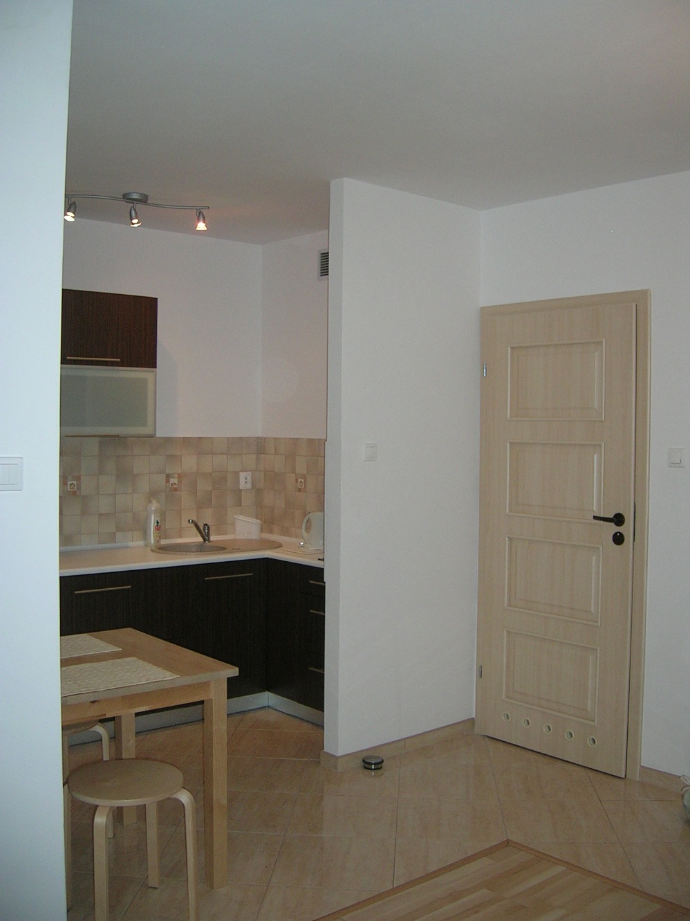 Wnętrze apartamentu: widok na aneks kuchenny z ciemnymi szafkami, beżowe płytki na ścianie i podłodze, drewniany stół z krzesłem oraz jasne drzwi z poziomymi panelami i okrągłymi wgłębieniami u dołu.