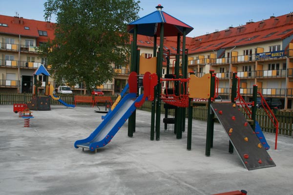 Kolorowy plac zabaw z wieżą, zjeżdżalnią i ścianką wspinaczkową na tle apartamentowca z balkonami.