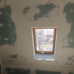 Krystian Barczyński - Poddasze ze skośnym sufitem w trakcie szpachlowania, widoczne ślady po łatach gipsowych. Okno dachowe z widokiem na rusztowanie. W tle wiszące kable.