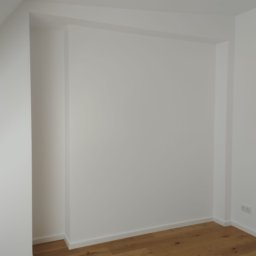 Krystian Barczyński - Białe, świeżo pomalowane ściany w rogu pokoju z drewnianą podłogą. Widoczne gniazdka elektryczne. Minimalistyczne wykończenie wnętrza.