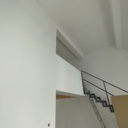 Krystian Barczyński - Białe, świeżo pomalowane wnętrze z metalowymi schodami i drabiną. Widoczne wykończenia sufitu i fragment otworu drzwiowego. Minimalistyczny design.