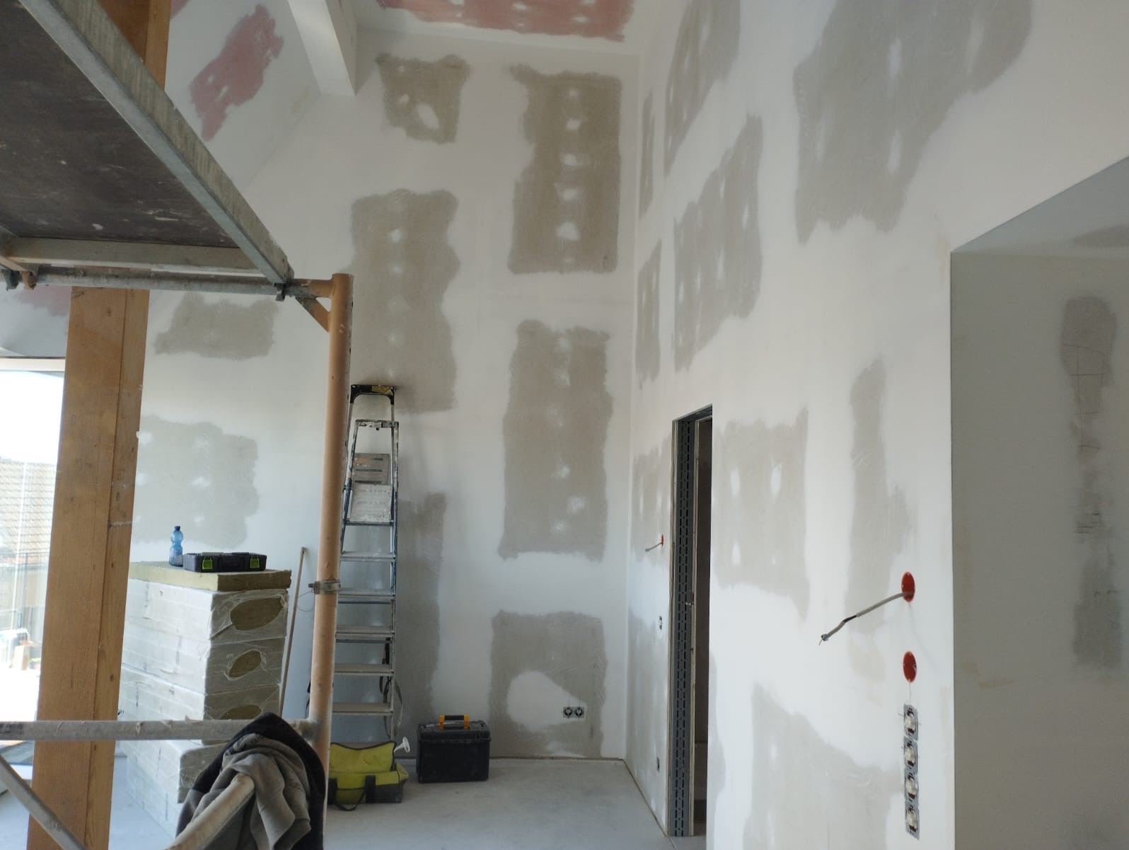 Wnętrze w trakcie szpachlowania, widoczne ściany z nałożoną warstwą szpachli, drabina, rusztowanie i narzędzia. Przygotowanie do dalszych prac wykończeniowych.