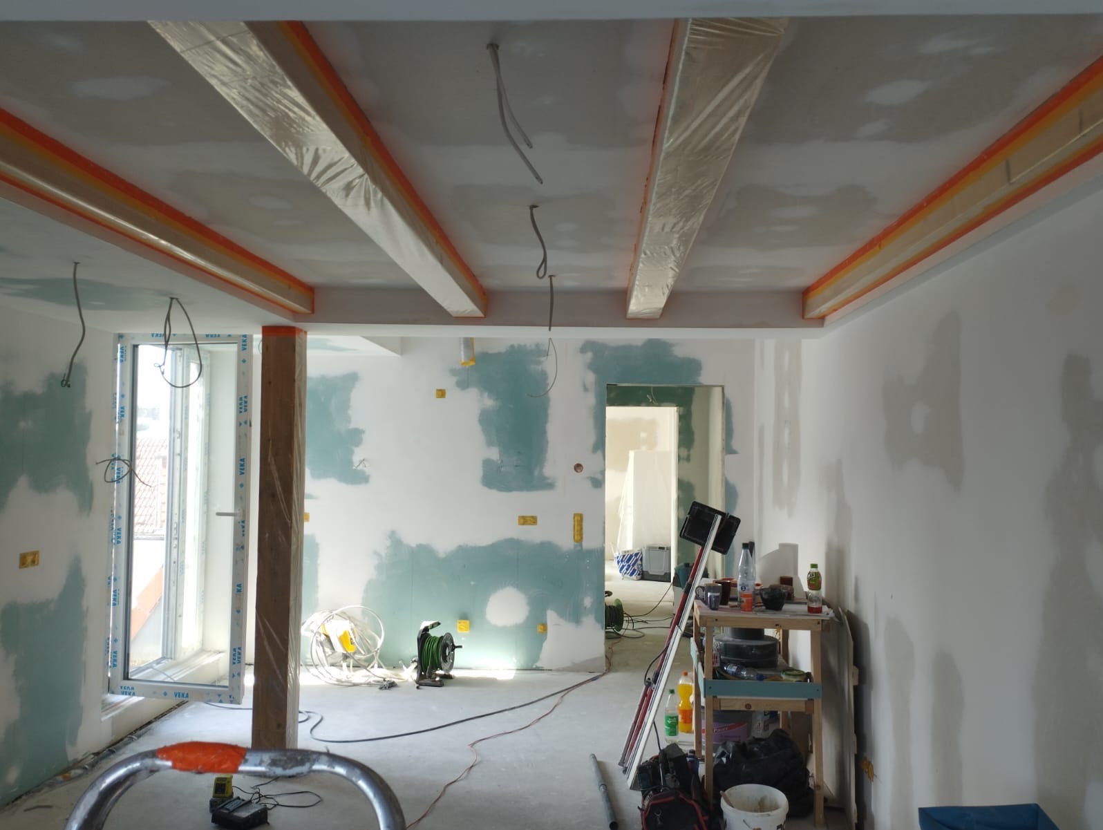 Wnętrze w trakcie prac: ściany z fragmentami szpachli, zabezpieczone folią belki sufitowe z pomarańczową taśmą, okno z profilem Veka, narzędzia i materiały budowlane.
