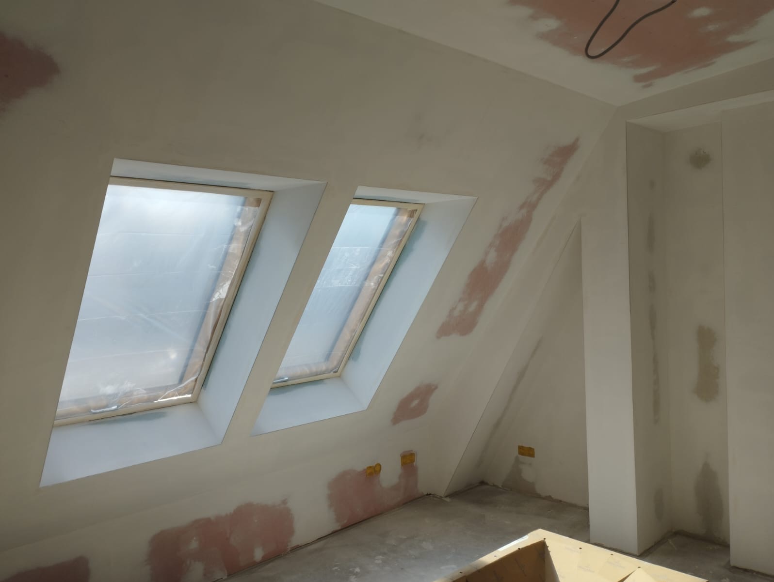 Poddasze po szpachlowaniu, z dwoma oknami dachowymi zabezpieczonymi folią. Ściany i sufit pokryte gładzią, widoczne różowe plamy po naprawach. Przygotowanie do malowania.