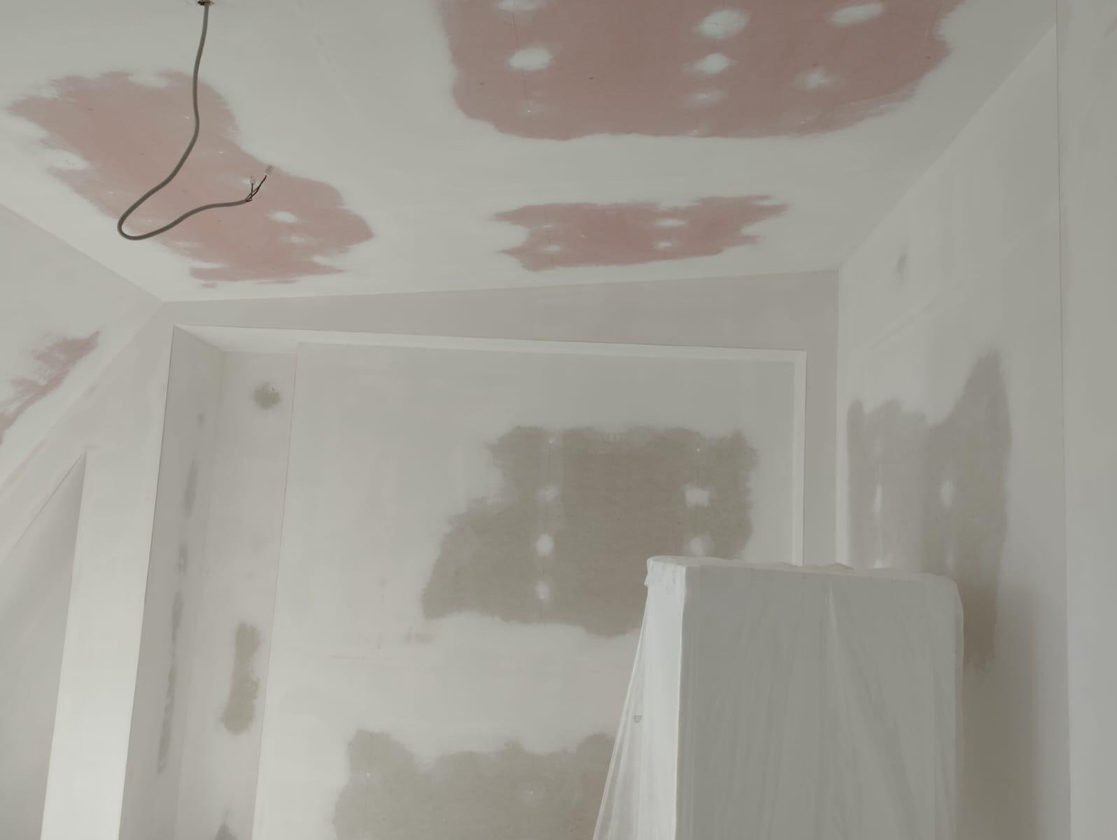 Pokój w trakcie prac wykończeniowych: ściany i sufit po szpachlowaniu, z widocznymi łatami i wystającym przewodem elektrycznym. Meble zabezpieczone folią.