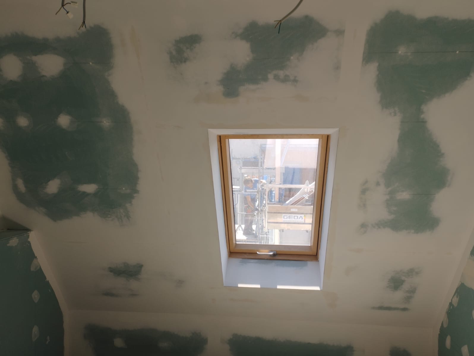 Poddasze ze skośnym sufitem w trakcie szpachlowania, widoczne ślady po łatach gipsowych. Okno dachowe z widokiem na rusztowanie. W tle wiszące kable.