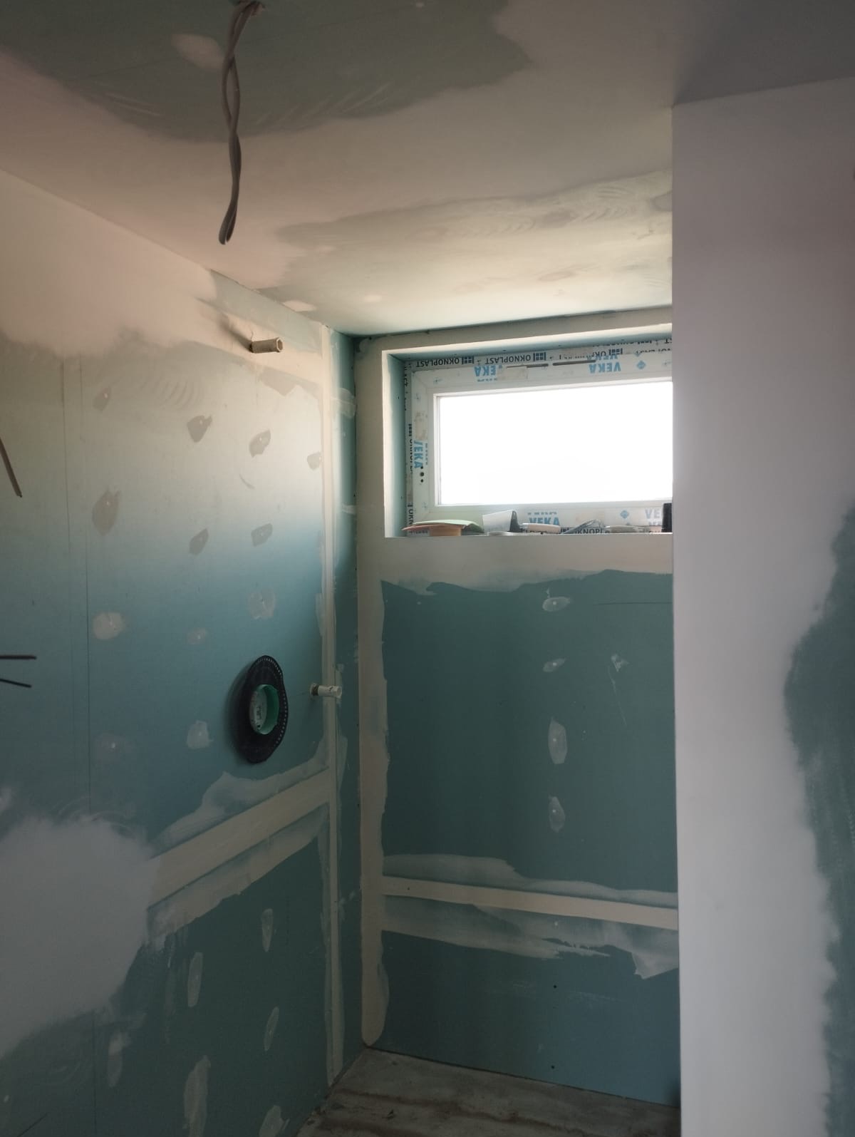 Narożnik łazienki w trakcie prac wykończeniowych: ściany z płyt gipsowo-kartonowych, widoczne łączenia, okno. W tle wystające przewody elektryczne.