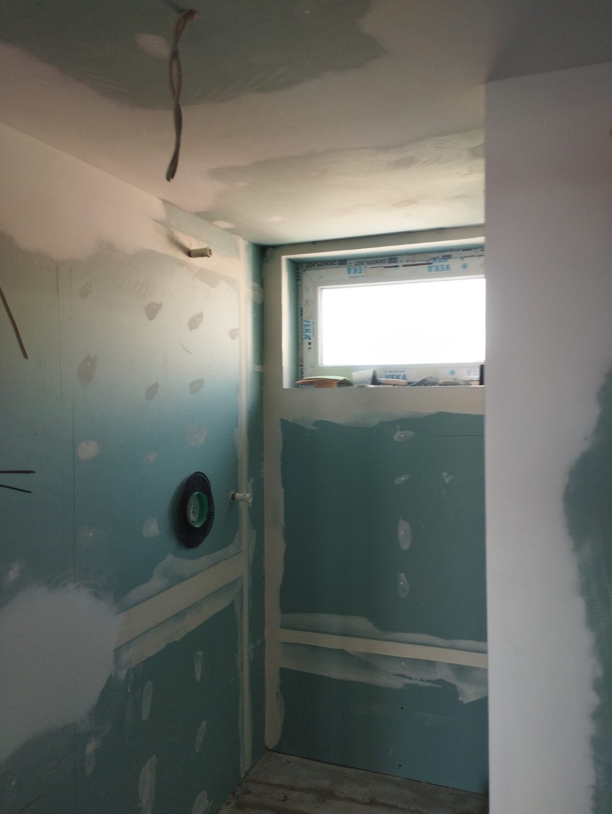 Wnętrze łazienki w trakcie prac wykończeniowych: ściany z płyt gipsowo-kartonowych, widoczne łączenia, okno z parapetem. Przygotowanie do dalszych etapów.