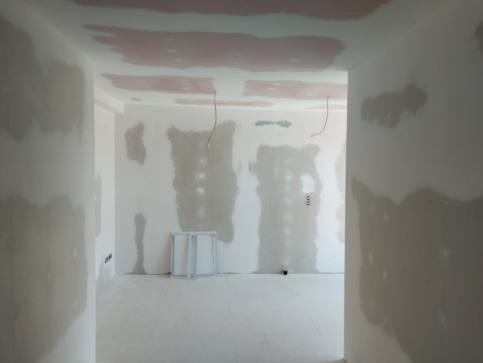 Wnętrze w trakcie szpachlowania: ściany i sufit pokryte warstwą gładzi, widoczne kable i rama drzwi. Przygotowanie do dalszych prac wykończeniowych.