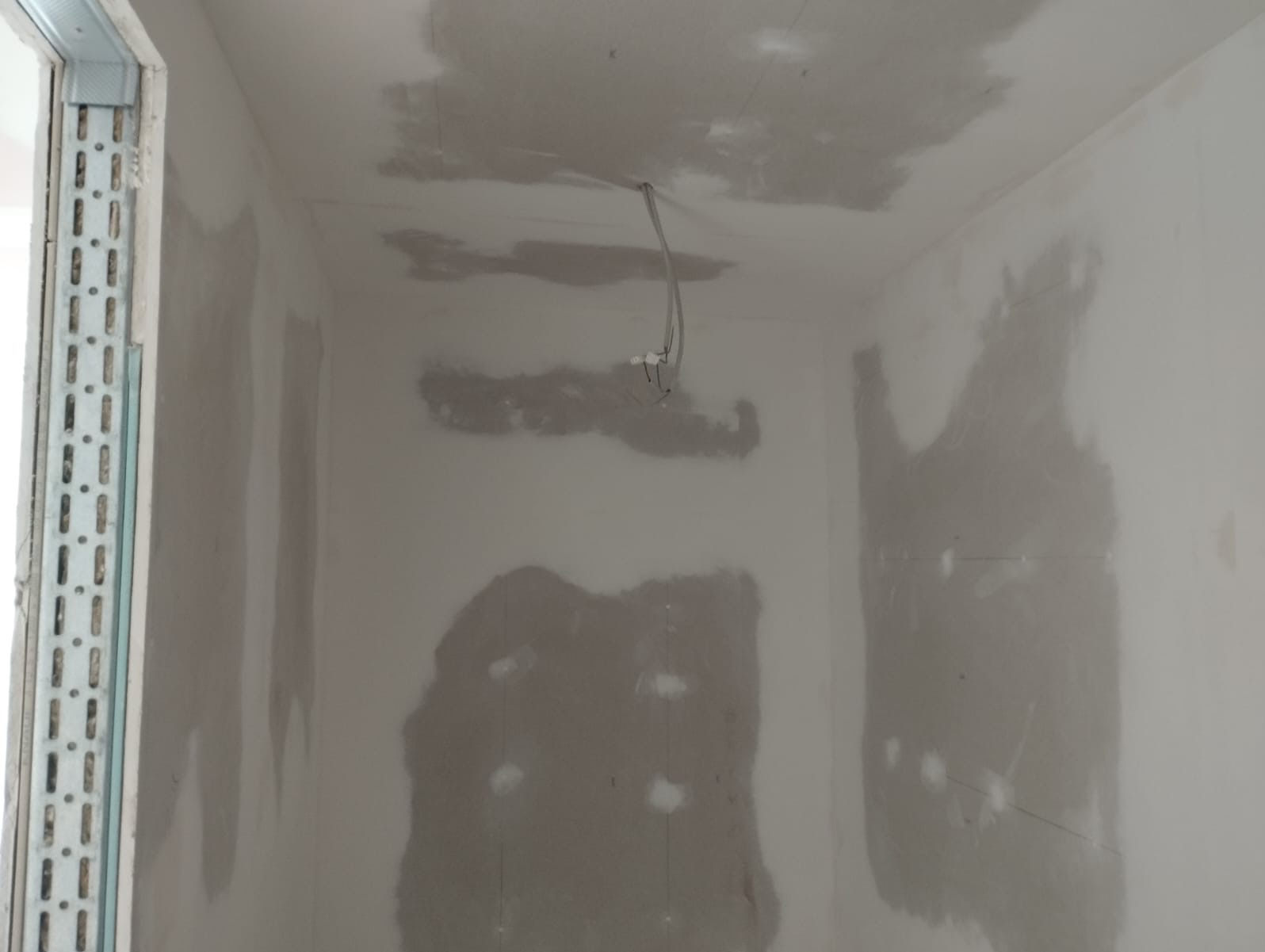 Wnętrze w trakcie szpachlowania: ściany i sufit pokryte szpachlą, widoczne profile przy ościeżnicy. Przewody elektryczne zwisają z sufitu.