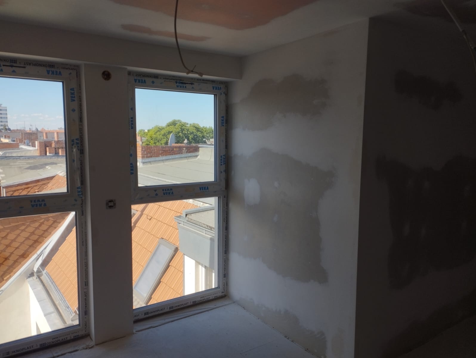 Ściana po szpachlowaniu, widoczne ślady po łatach gipsowych. Okna PCV z widokiem na dachy budynków. Etap prac wykończeniowych w nowym mieszkaniu.