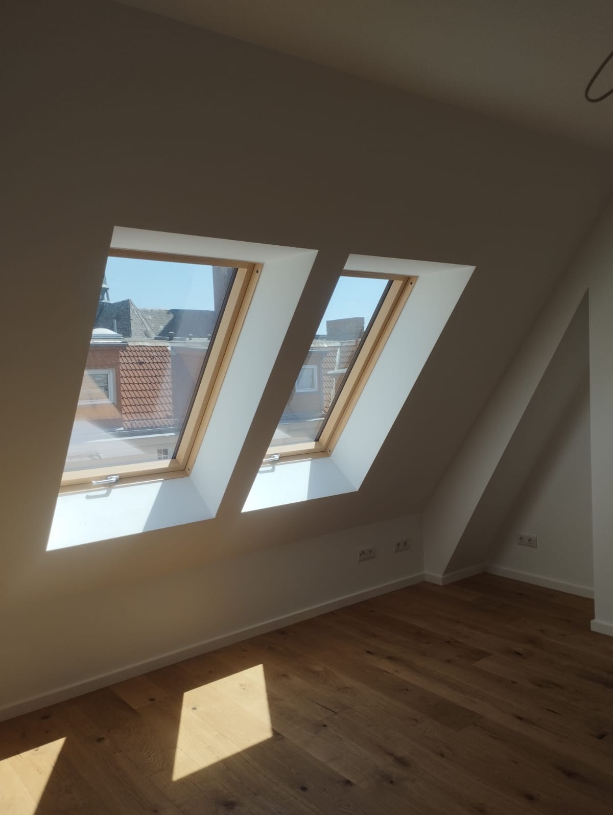 Jasne poddasze po remoncie: dwa okna dachowe z drewnianą ramą, parkiet, świeżo pomalowane białe ściany i gniazdka elektryczne. Widok na dachy budynków przez okna.
