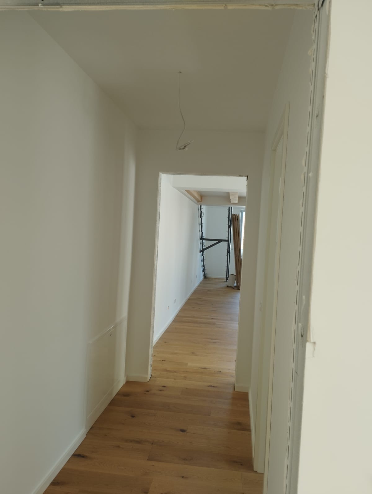 Jasny korytarz po remoncie z drewnianą podłogą. Widoczne ściany pomalowane na biało, a w głębi pomieszczenia rusztowanie i deski.