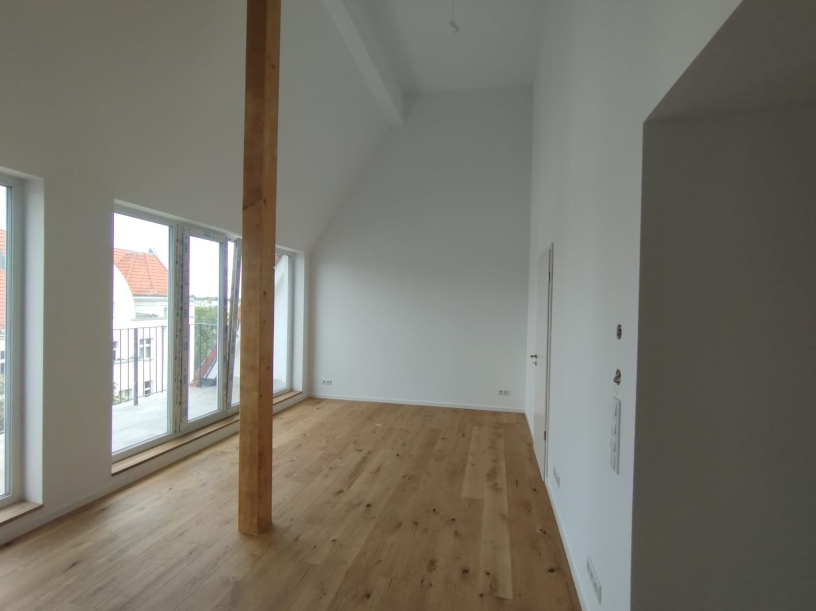 Jasne, przestronne wnętrze po remoncie: drewniana podłoga, białe ściany, duże okna z widokiem na zewnątrz. Widoczna drewniana belka konstrukcyjna.