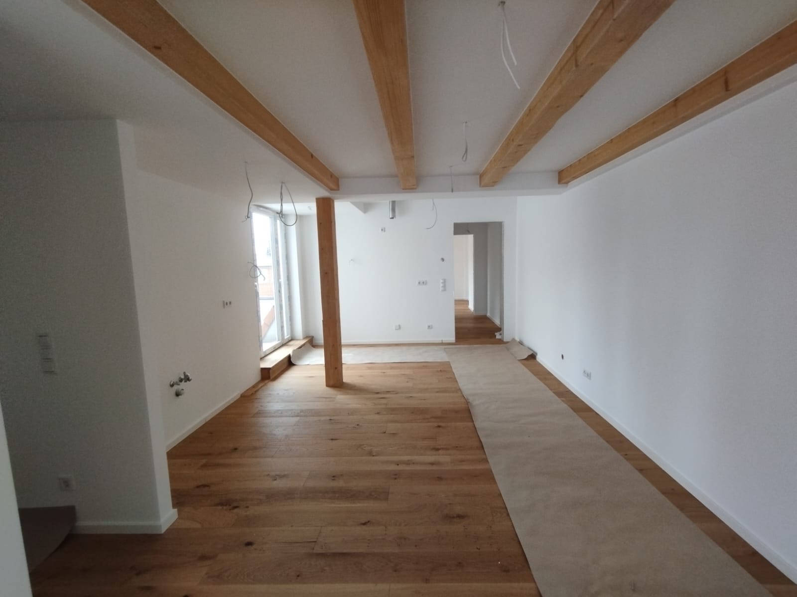 Jasne wnętrze w trakcie wykańczania: drewniana podłoga, białe ściany, surowe belki stropowe. Podłoga częściowo zabezpieczona kartonem.