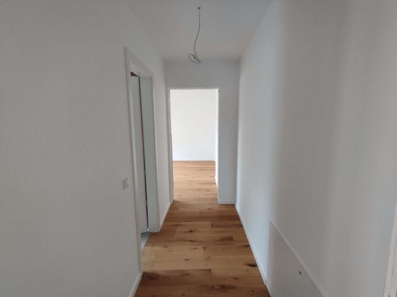 Jasny korytarz po remoncie: białe ściany, drewniana podłoga, widoczne ościeżnice drzwiowe. Kabel zwisa z sufitu. Perspektywa wnętrza.