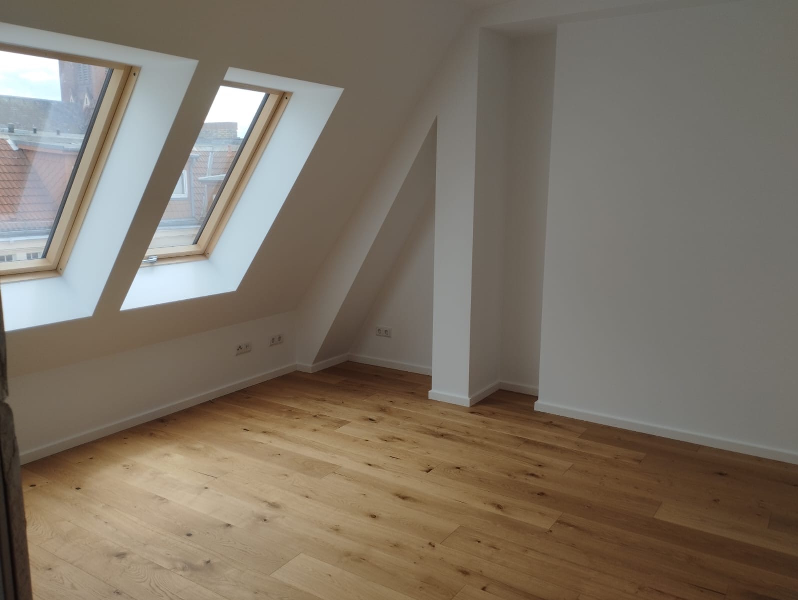 Jasne, wykończone poddasze z drewnianą podłogą, białymi ścianami i dwoma oknami dachowymi. Widoczne gniazdka elektryczne.