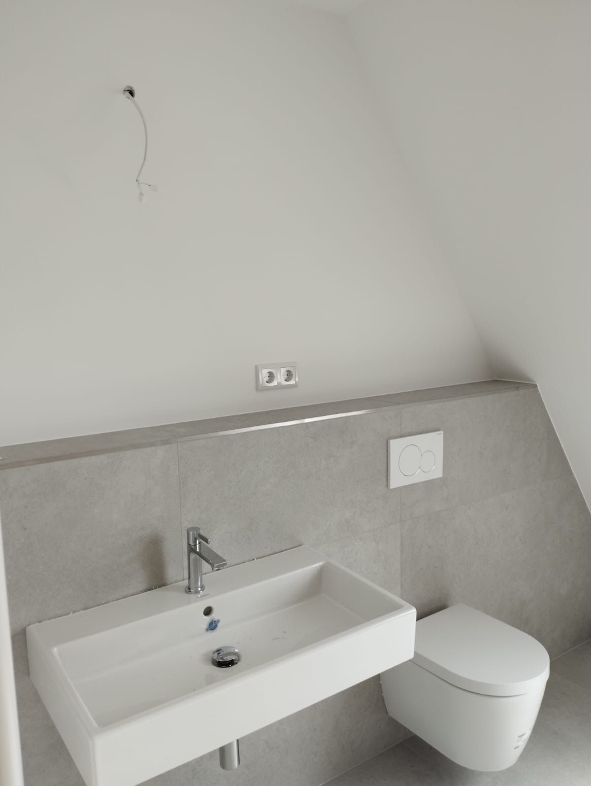 Nowoczesna łazienka na poddaszu z umywalką, toaletą, szarą glazurą i wystającą instalacją elektryczną. Minimalistyczny design, jasne kolory.