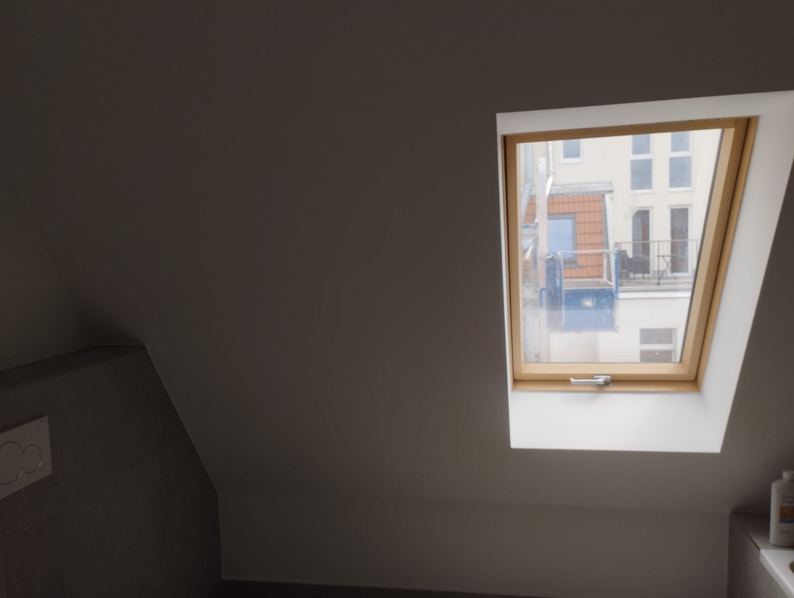 Szare poddasze z oknem dachowym i widokiem na budynek z rusztowaniem. Świeżo pomalowane ściany. Minimalistyczny styl.