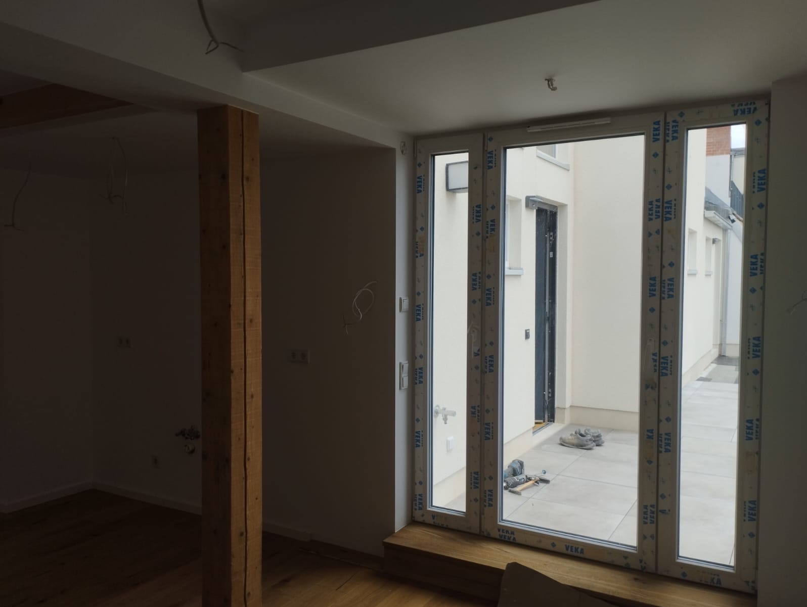 Wnętrze w trakcie remontu: drewniany słup, nowe okno balkonowe z widokiem na patio, narzędzia na podłodze. Widoczne przewody elektryczne i elementy drewniane.