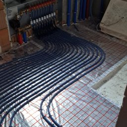 DAW-MON MONIKA WALKOWIAK - Niebieskie rury ogrzewania podłogowego ułożone na folii z czerwoną siatką, widoczne elementy rozdzielacza i piony instalacyjne przy ścianie w surowym stanie.