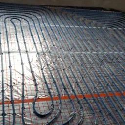 Ułożona instalacja ogrzewania podłogowego z niebieskich rurek na folii z czerwoną siatką, widok z góry, surowy stan wykończenia wnętrza.