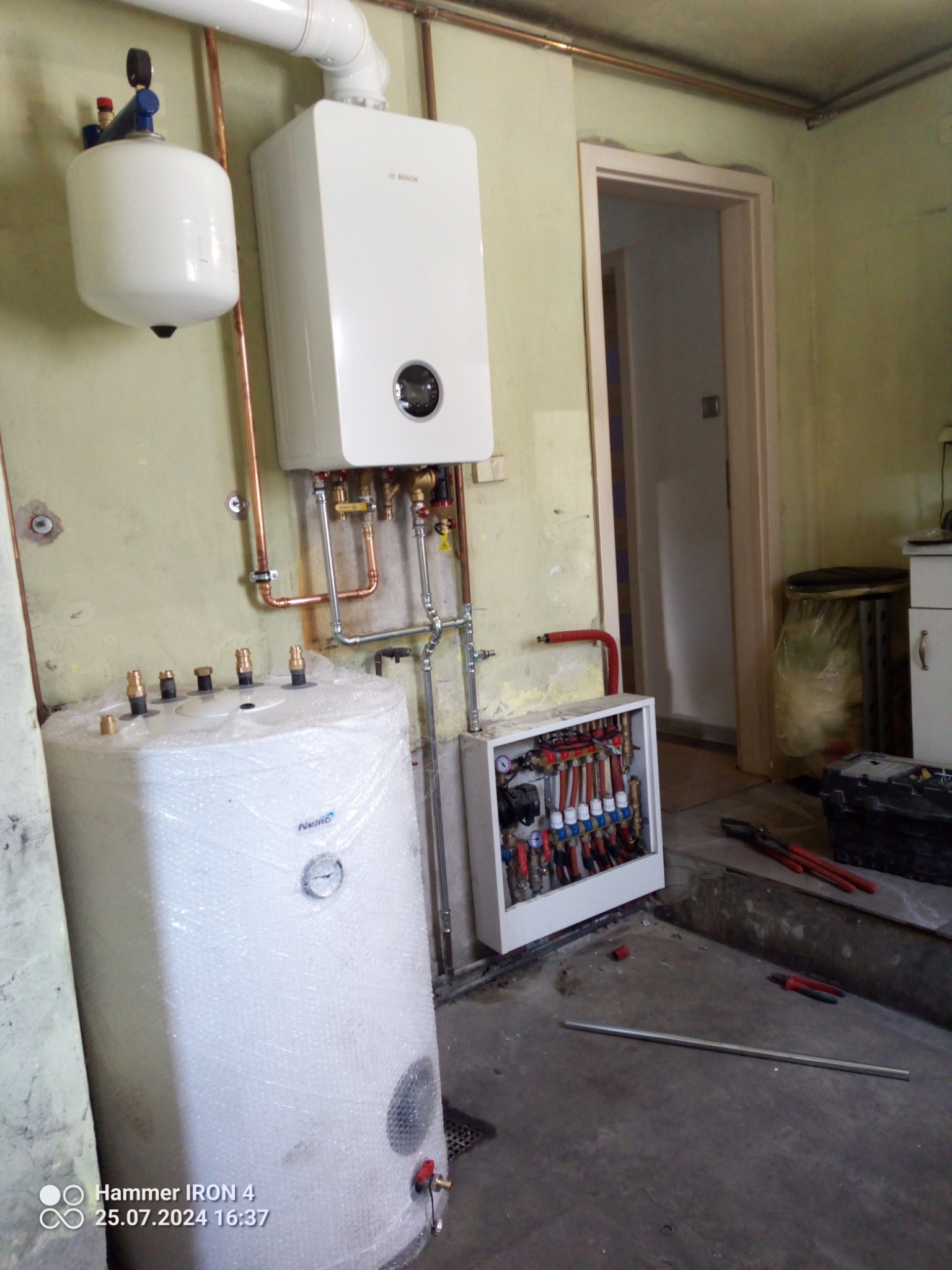 Biały piec gazowy Bosch, zbiornik Nemo owinięty folią bąbelkową i rozdzielacz instalacji grzewczej z rurami. Widoczne miedziane rury i narzędzia hydrauliczne.