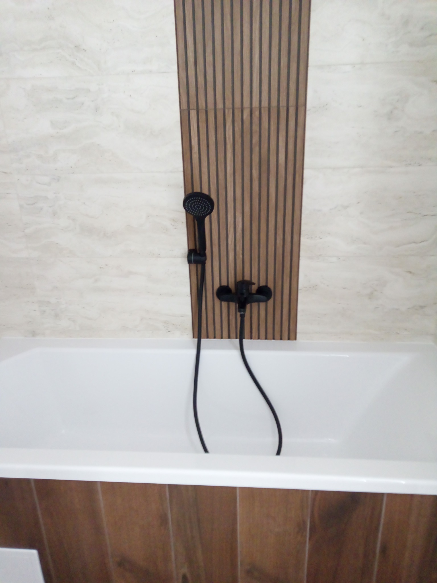 Nowoczesna, czarna armatura natryskowa zamontowana nad białą wanną, na tle beżowych płytek i dekoracyjnej drewnianej listwy. Minimalistyczny design łazienki.