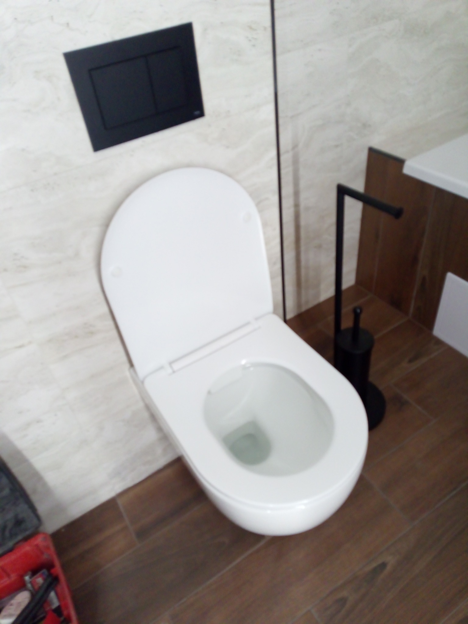 Nowoczesna toaleta wisząca z czarnym przyciskiem spłukującym i szczotką WC. Brązowe płytki podłogowe i beżowe ścienne tworzą spójną całość.