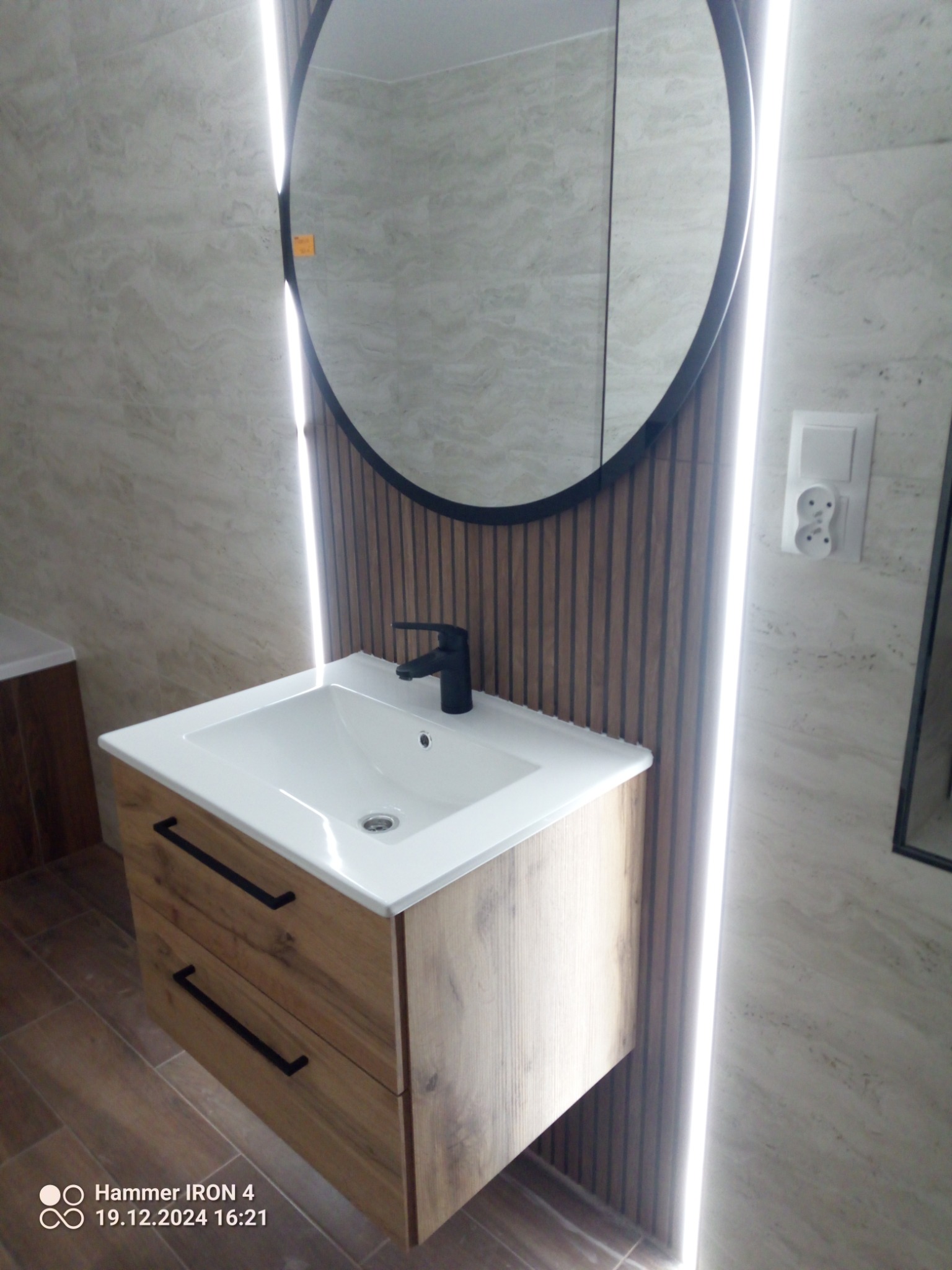 Nowoczesna łazienka: szafka z umywalką i czarną armaturą, okrągłe lustro z podświetleniem LED na tle płytek imitujących drewno i beżowych ścian.