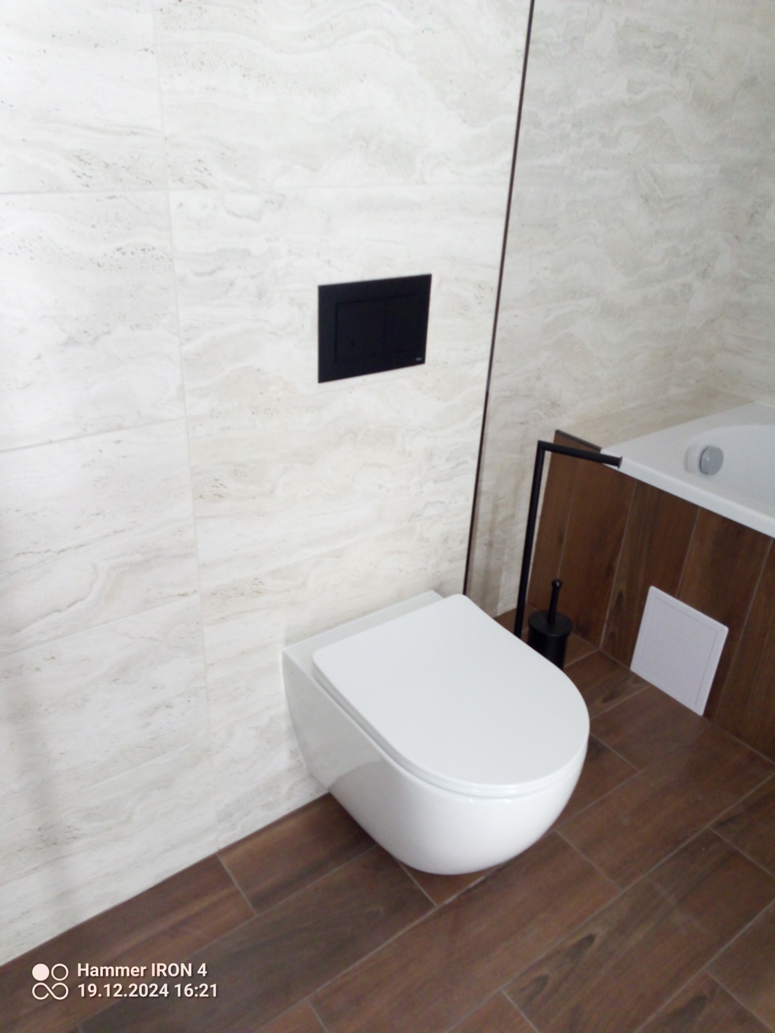 Nowoczesna toaleta podwieszana z czarnym panelem sterującym na tle marmurowej ściany i drewnopodobnej podłogi w wykończonej łazience.